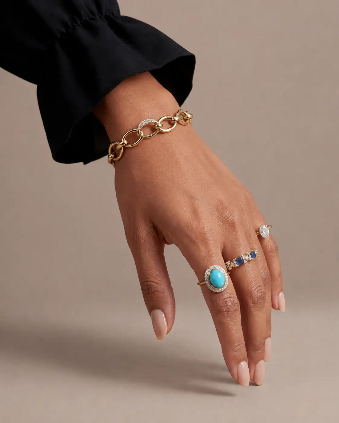 A close-up of a hand wearing a gold chain bracelet and several gold rings, including one with a turquoise oval stone and others with blue and clear stones, against a neutral beige background.