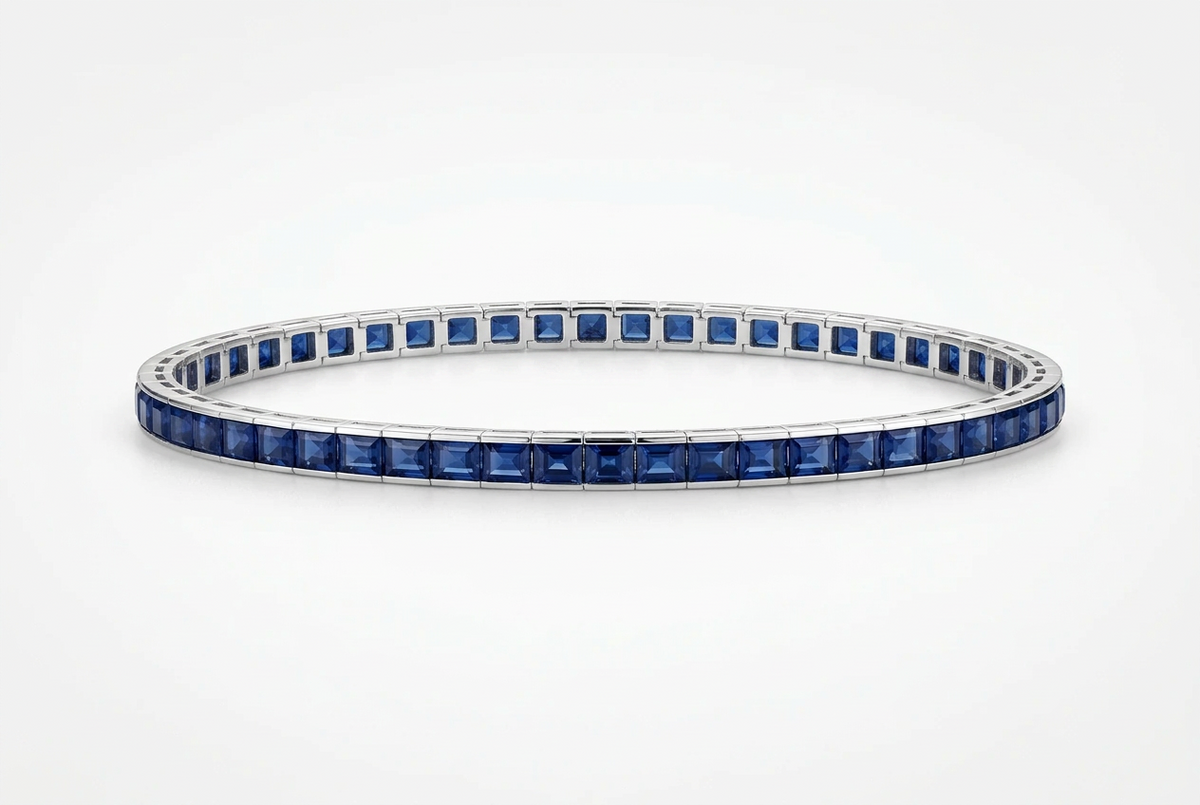 A tennis bracelet featuring square-cut deep blue sapphires set in a polished silver-colored metal, shown against a clean white background.